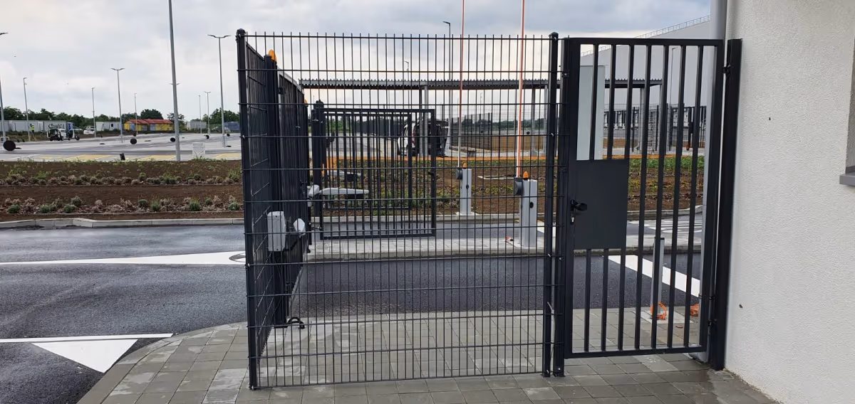 Metalna ograda i kapija sa parking rampama u pozadini.